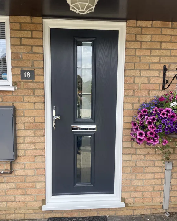 Anthracite grey composite front door with two slim vertical glass panels and chrome furniture, in a white frame on a buff brick house.