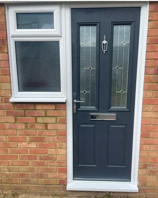 Anthracite grey composite front door with bevelled glass panels beside a white uPVC window with obscure glass, in a red brick wall.