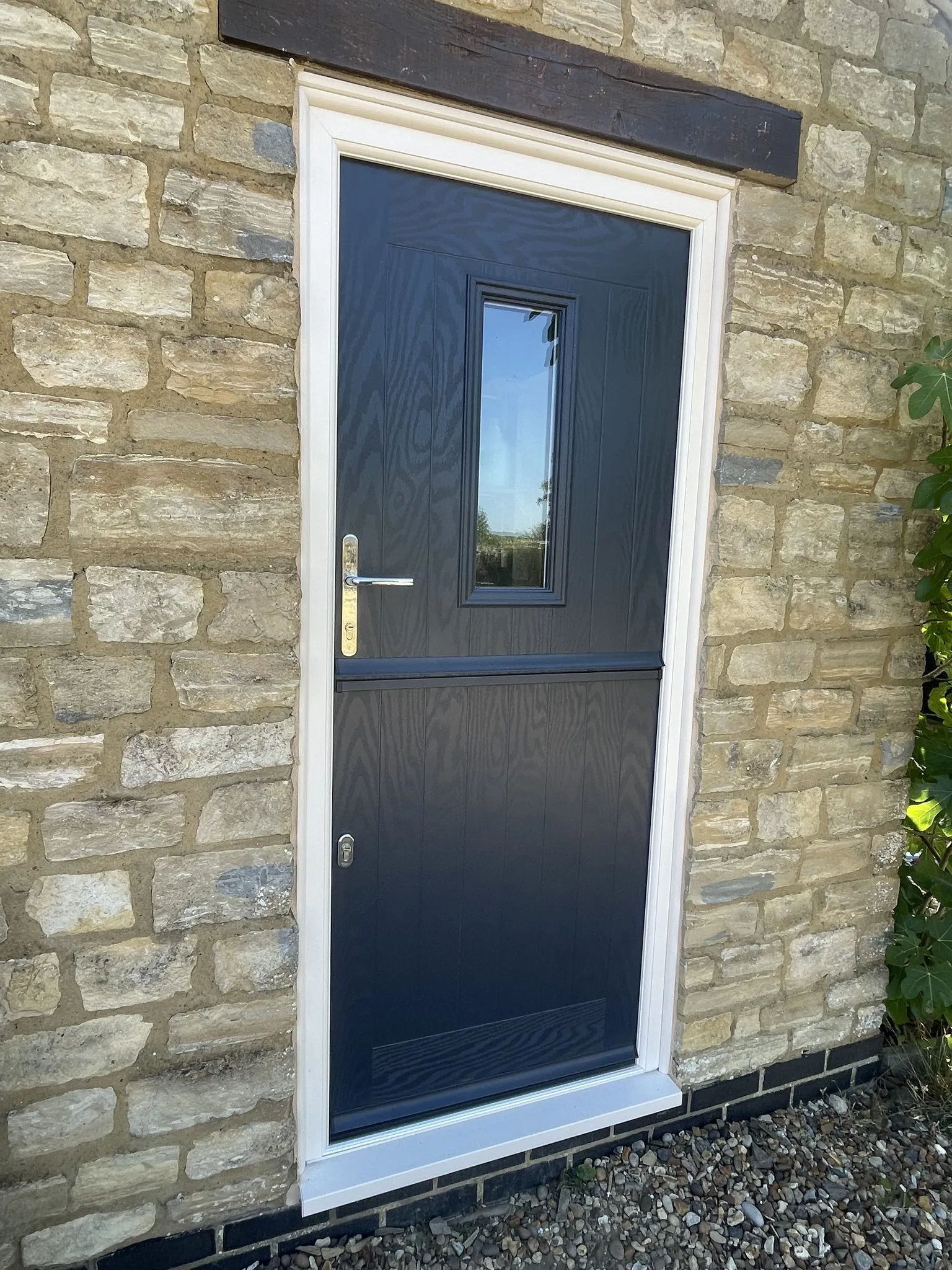 Anthracite grey composite stable door with a small glazed panel, in a cream frame set into a stone cottage wall under a timber lintel.