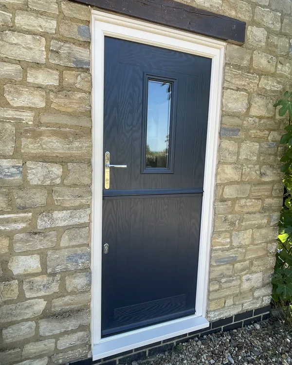 Anthracite grey composite stable door with a small glazed panel, in a cream frame set into a stone cottage wall under a timber lintel.