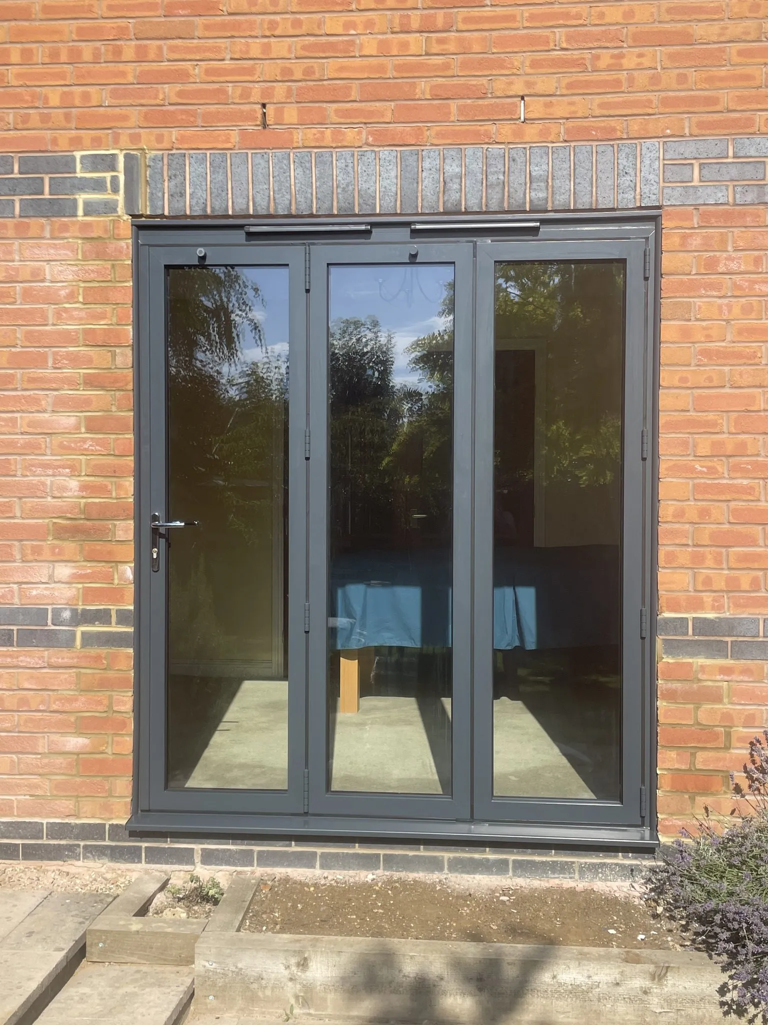 Three-panel anthracite grey bi-fold doors fitted in a red brick wall.