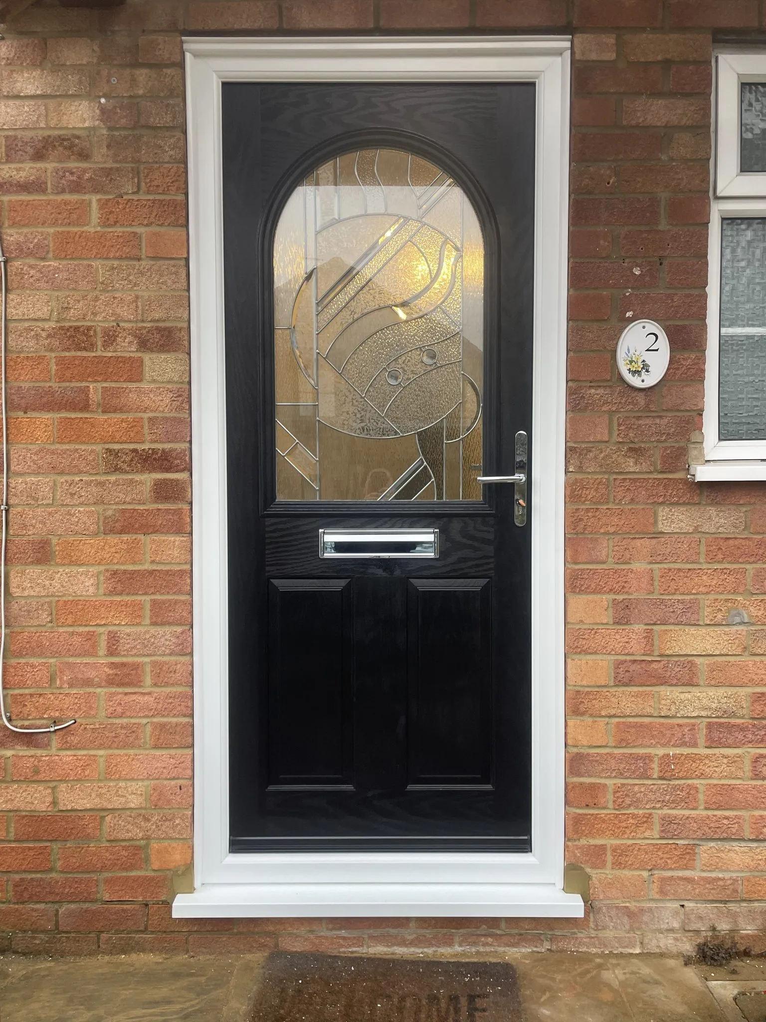 Black composite front door with a large arched decorative glass panel and chrome furniture, in a white frame on a red brick house.