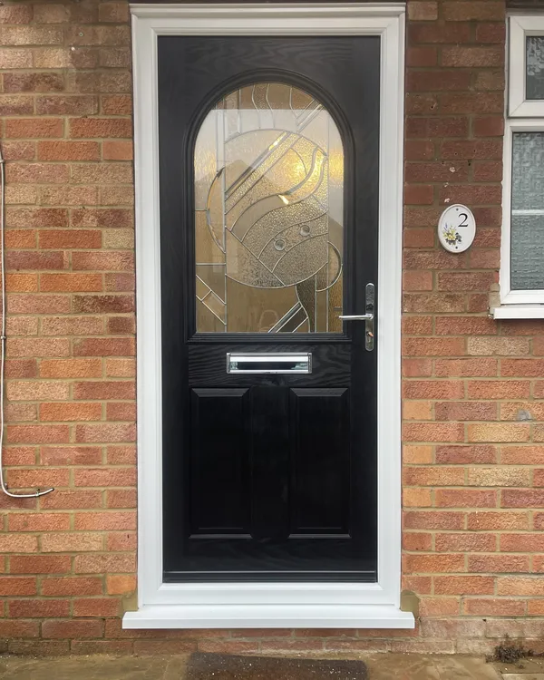 Black composite front door with a large arched decorative glass panel and chrome furniture, in a white frame on a red brick house.
