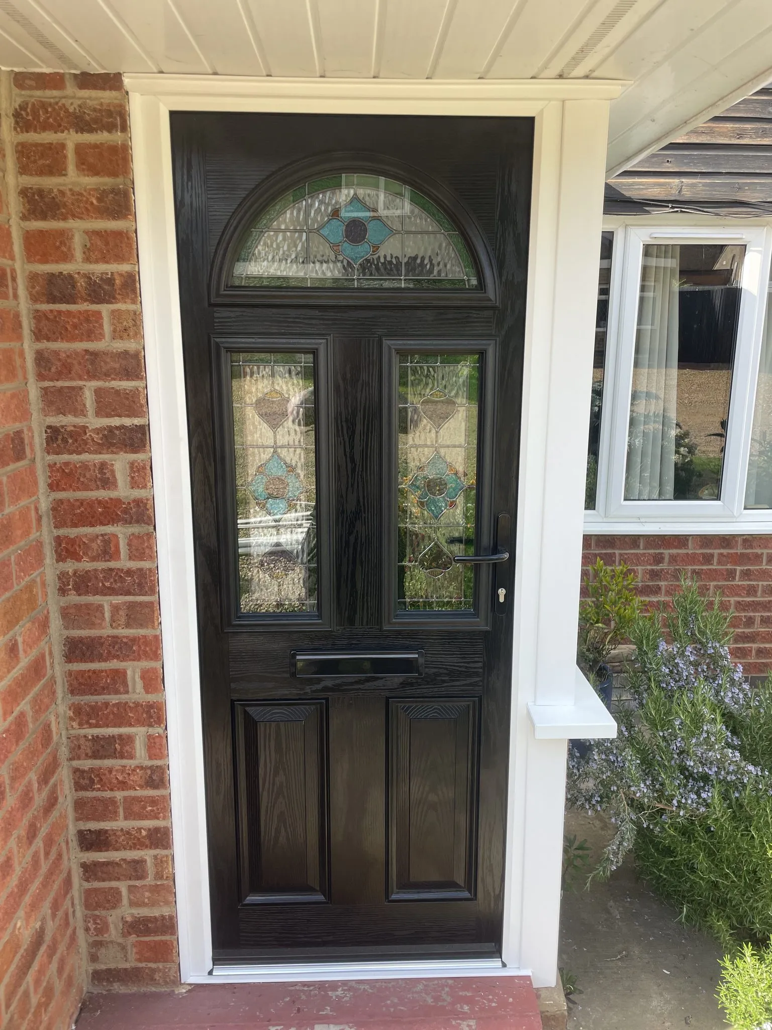 Black woodgrain composite front door with an arched stained glass top panel and matching glazed panels, beside a white uPVC porch window.