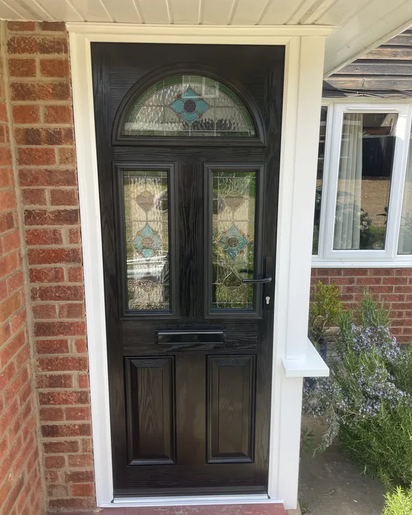 Black woodgrain composite front door with an arched stained glass top panel and matching glazed panels, beside a white uPVC porch window.