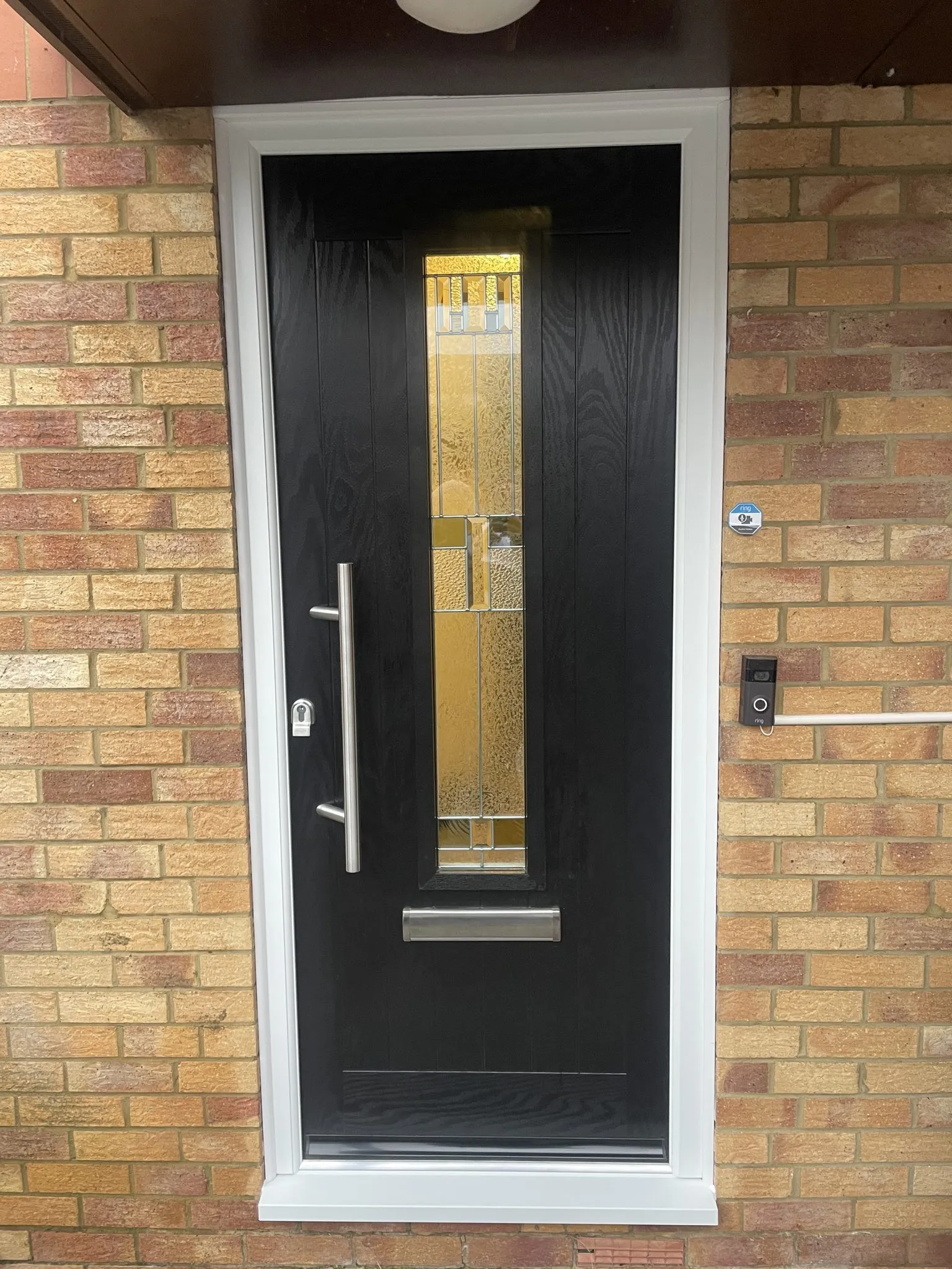 Contemporary black composite front door with a tall decorative glass strip and stainless steel bar handle, in a white frame on a buff brick house.