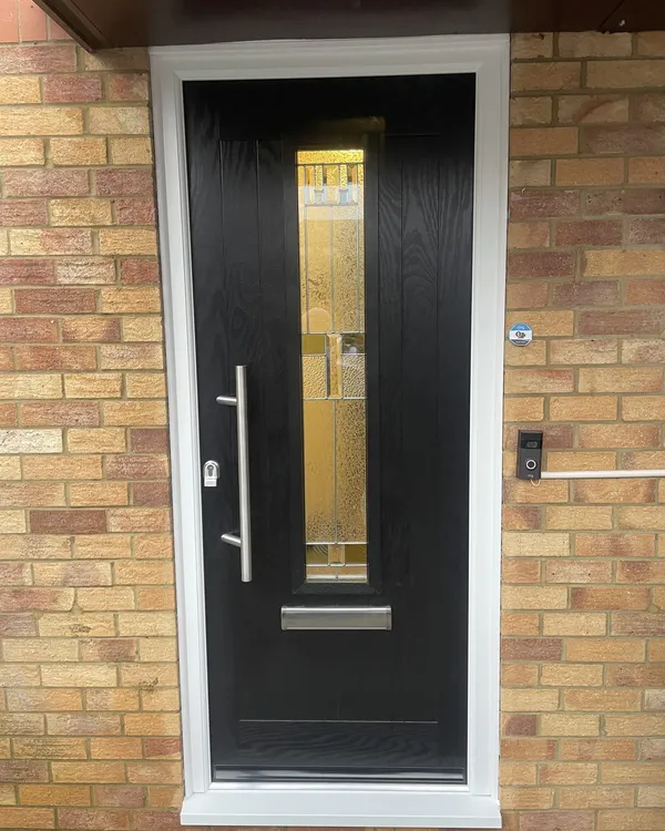 Contemporary black composite front door with a tall decorative glass strip and stainless steel bar handle, in a white frame on a buff brick house.