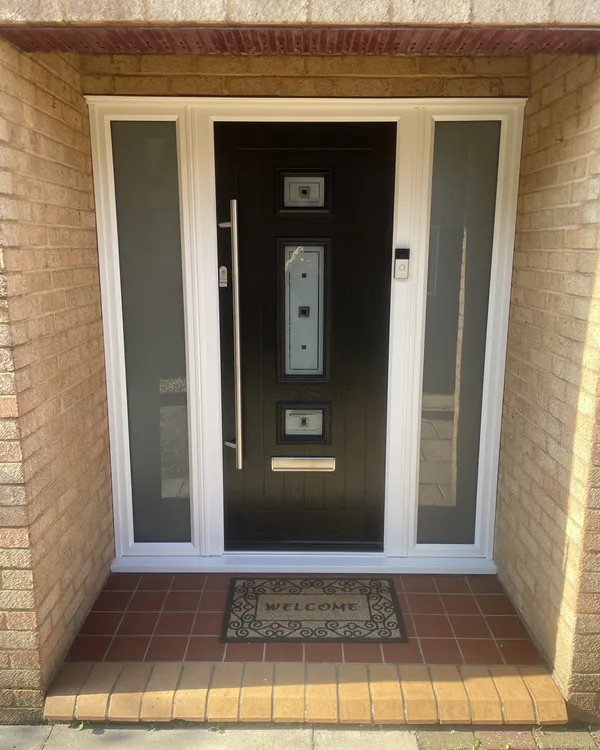 Contemporary black composite front door with a long chrome bar handle and glazed inserts, flanked by frosted full-height side panels in a recessed brick porch.