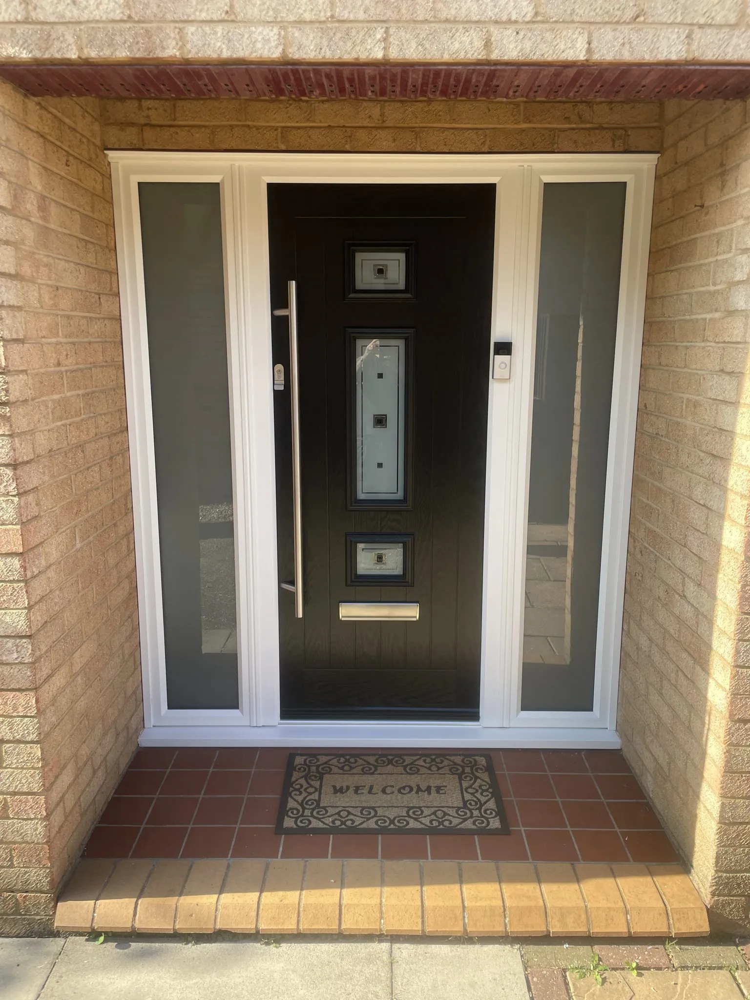 Contemporary black composite front door with a long chrome bar handle and glazed inserts, flanked by frosted full-height side panels in a recessed brick porch.