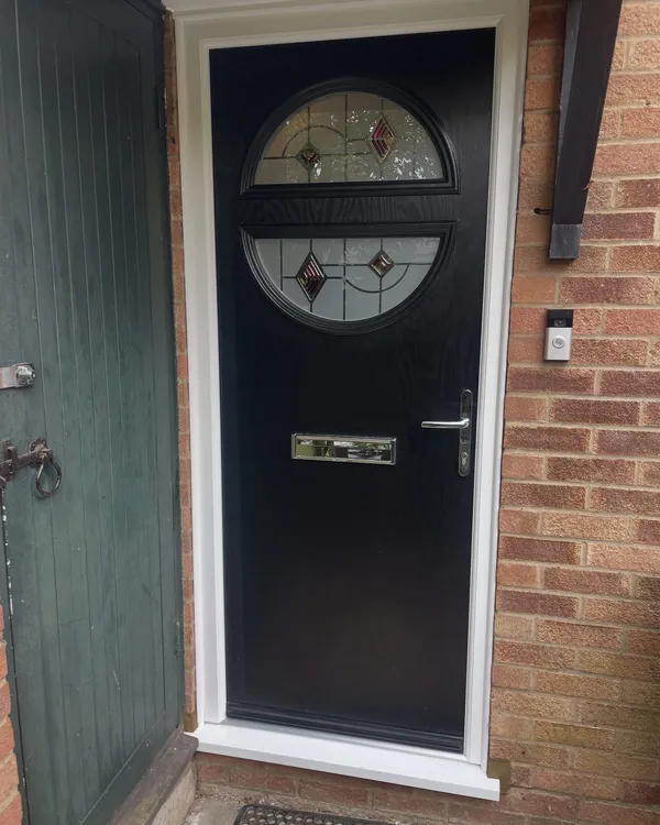 Black composite front door with a circular decorative glazed panel split by a central rail, in a white frame on a brick house.