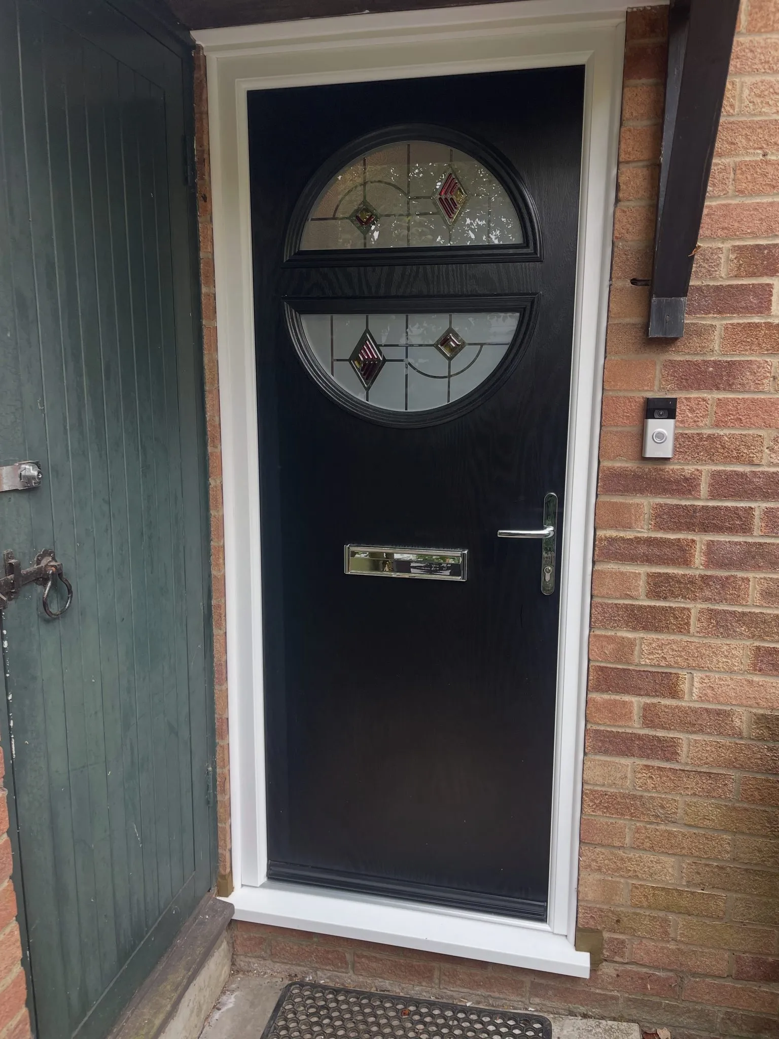 Black composite front door with a circular decorative glazed panel split by a central rail, in a white frame on a brick house.