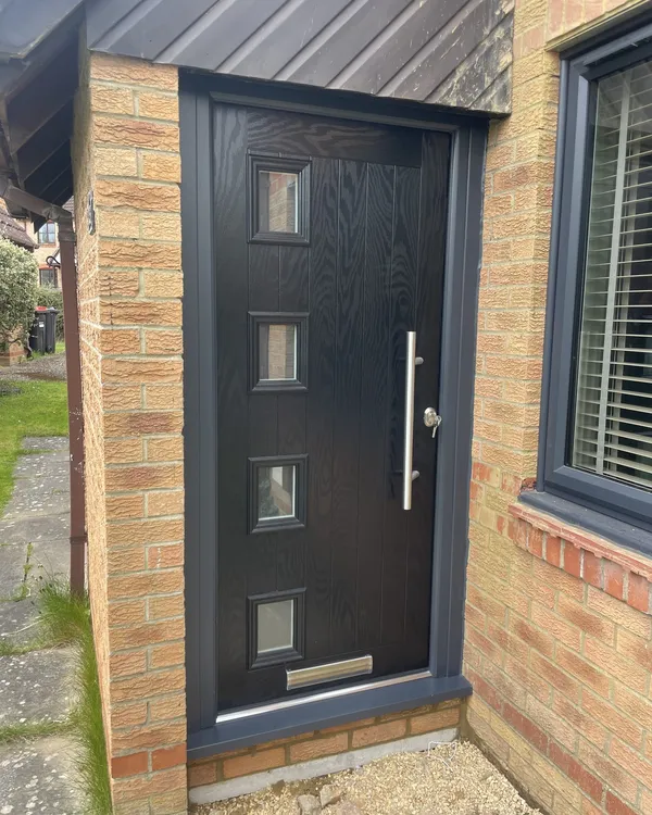 Contemporary black composite front door with four small square glazed panels and a chrome bar handle, in an anthracite grey frame beside a matching grey window.