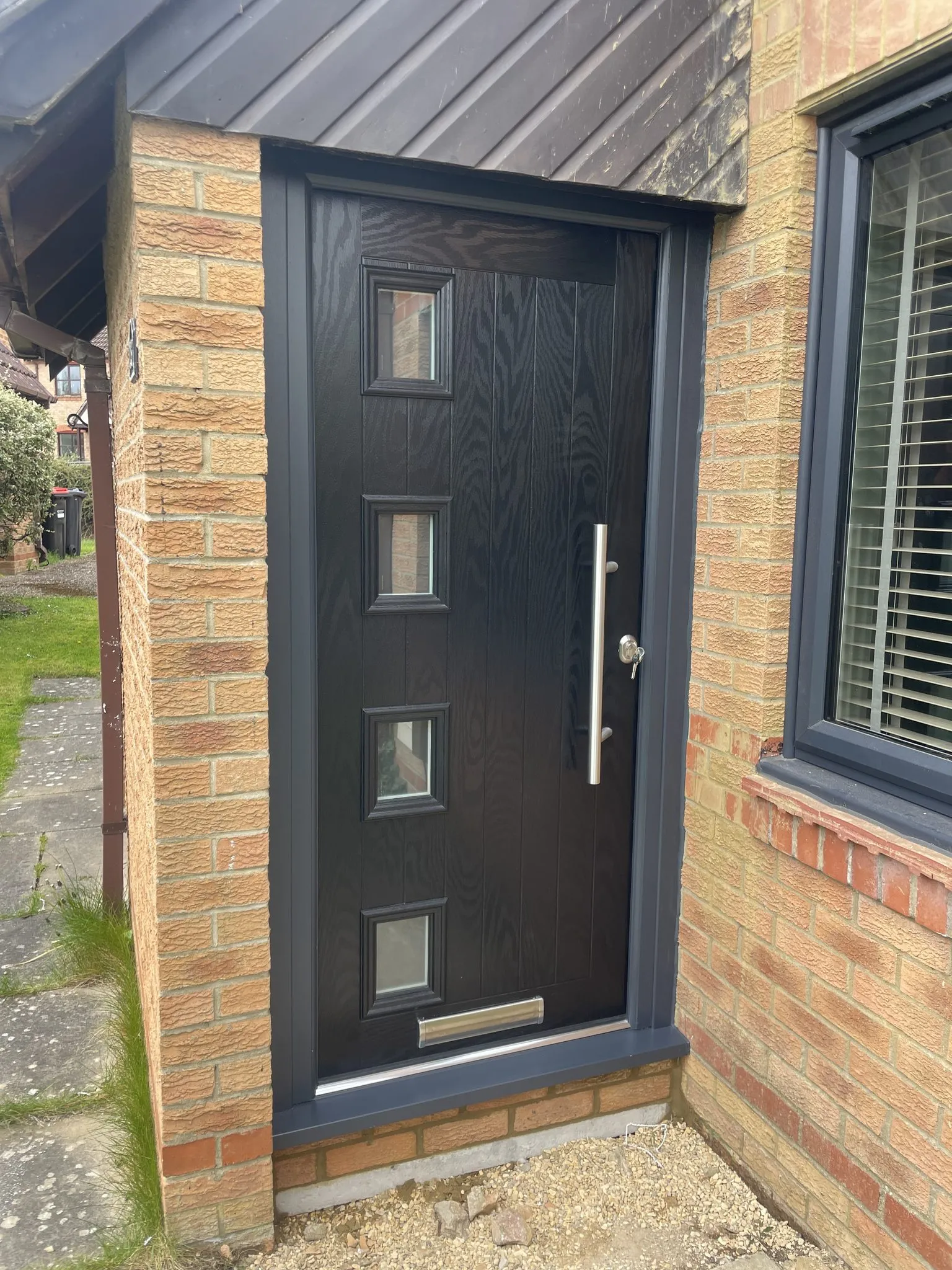 Contemporary black composite front door with four small square glazed panels and a chrome bar handle, in an anthracite grey frame beside a matching grey window.