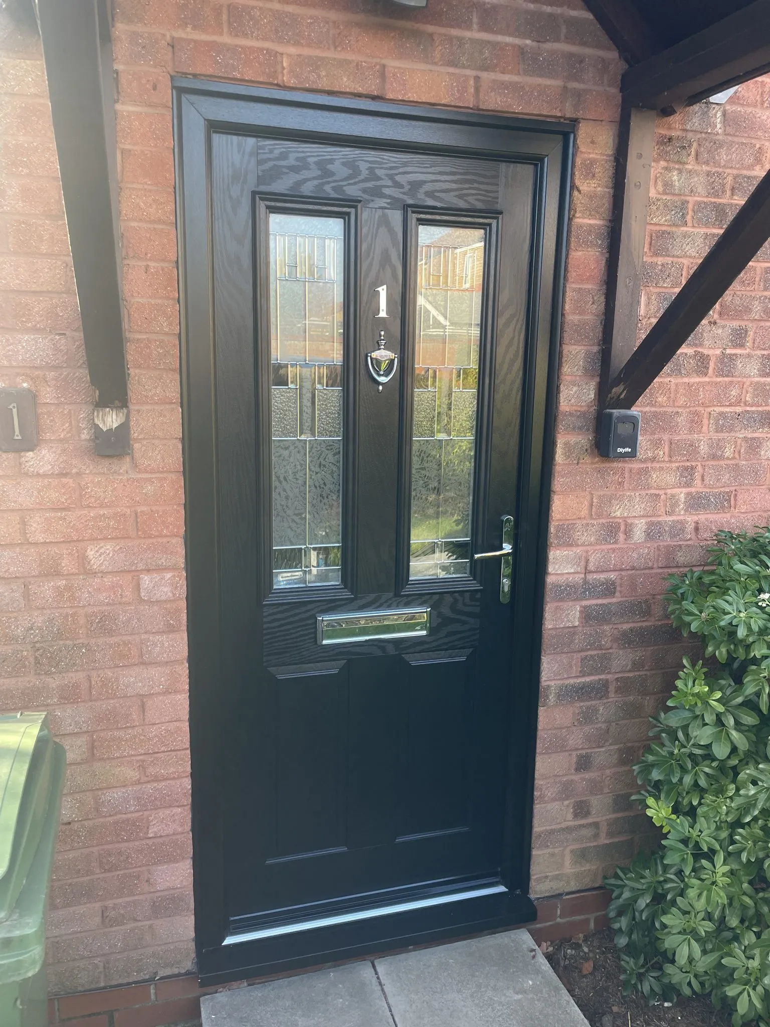 Black woodgrain composite front door with glazed panels, chrome numeral and knocker, in a black frame below a timber canopy.