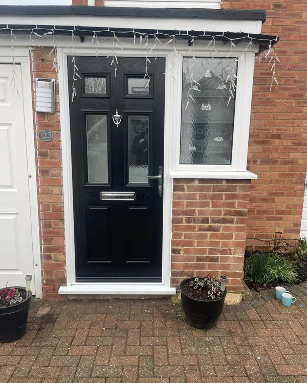 Black composite front door with four glazed panels and chrome furniture beside a white uPVC side window, on a brick house.