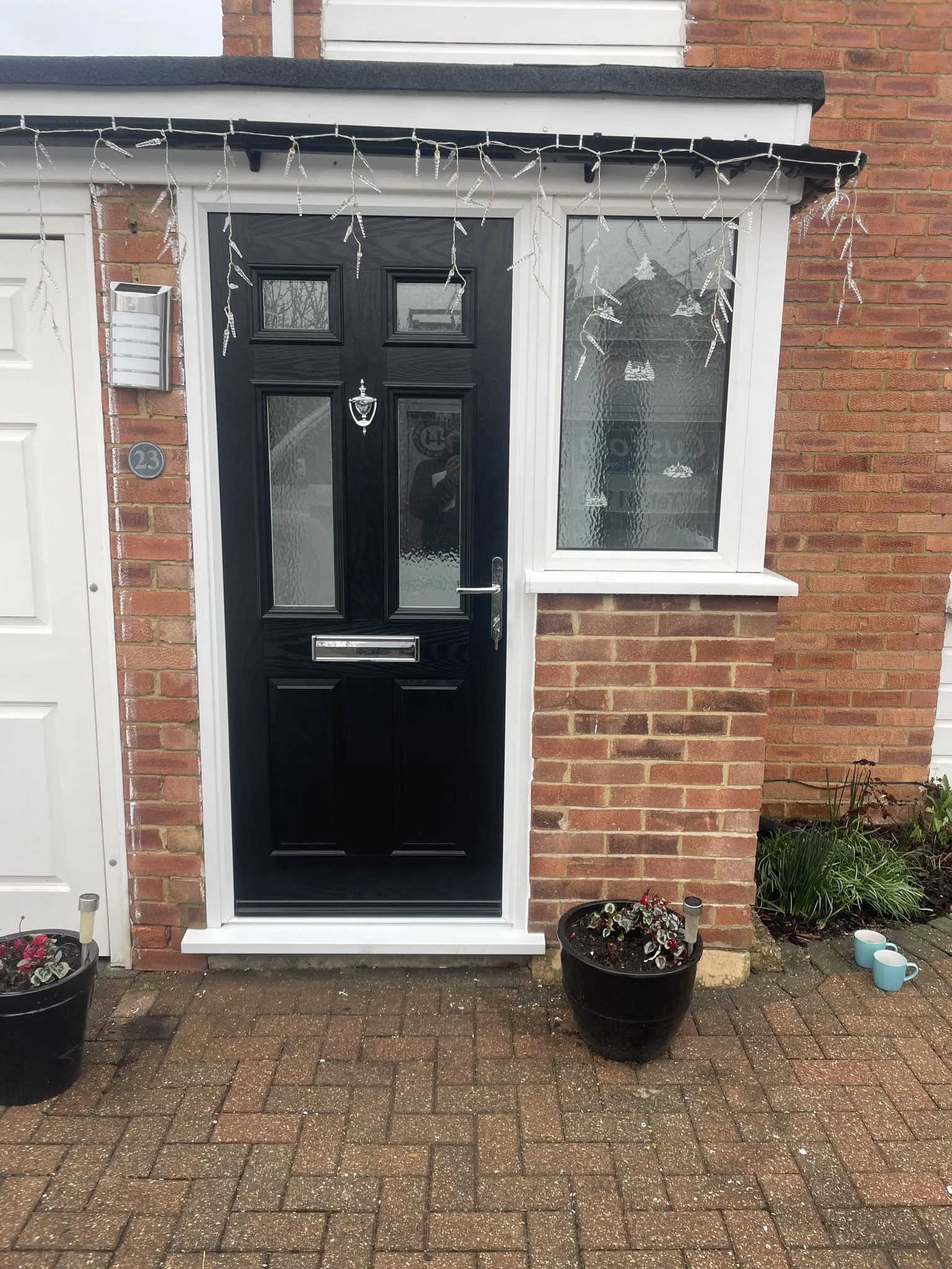 Black composite front door with four glazed panels and chrome furniture beside a white uPVC side window, on a brick house.
