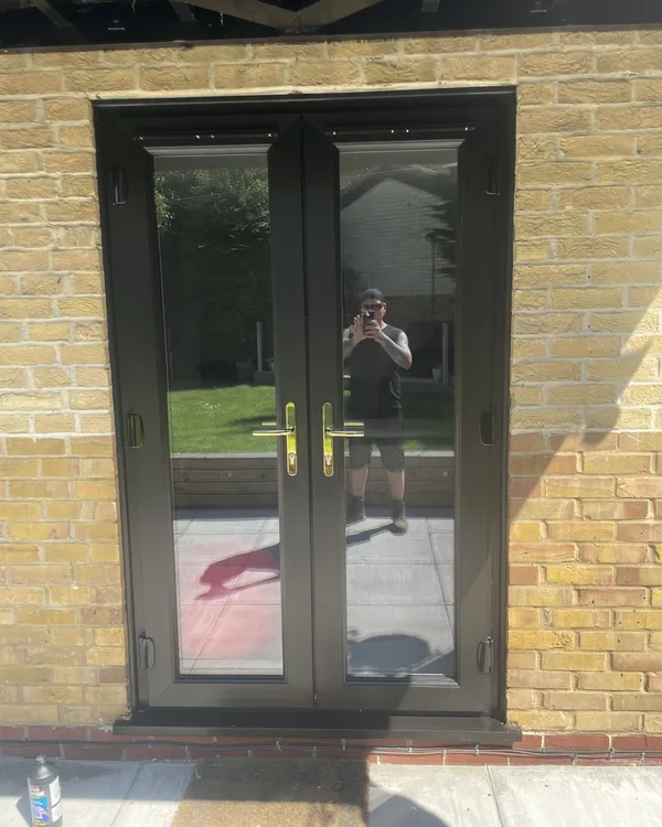Black uPVC French doors with gold handles fitted in a buff brick wall, opening onto a patio.