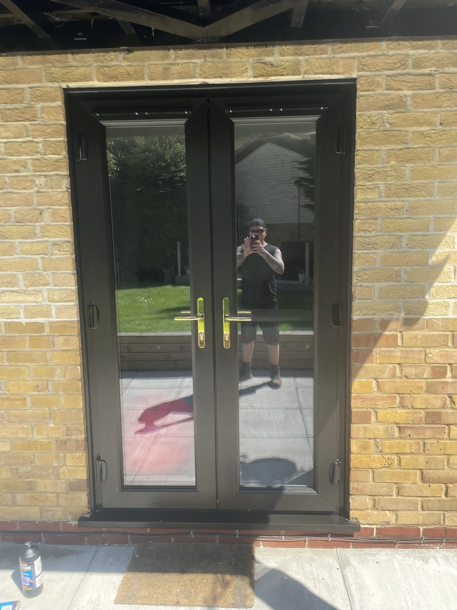 Black uPVC French doors with gold handles fitted in a buff brick wall, opening onto a patio.