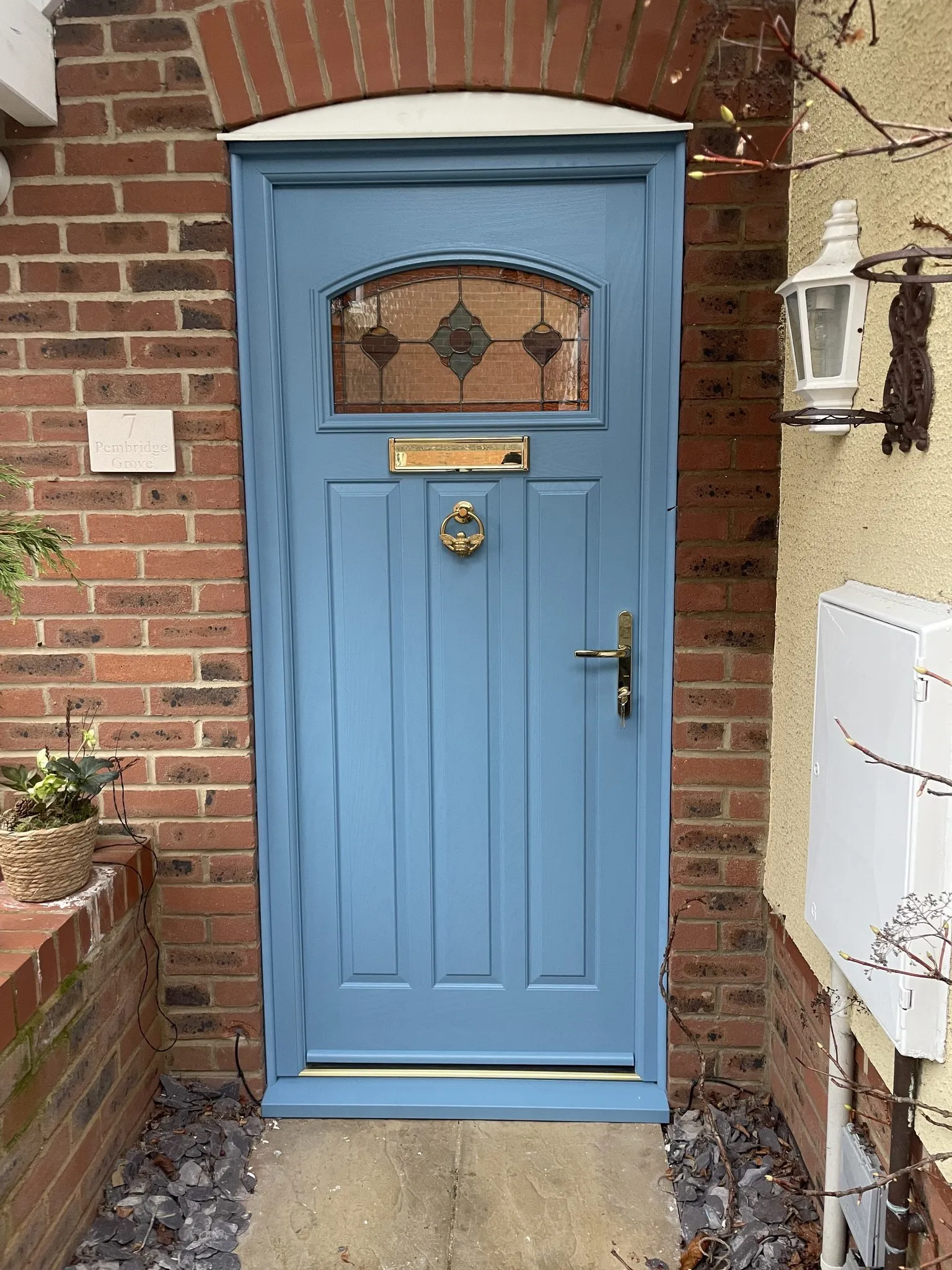 Mid blue cottage-style composite door with a stained glass top panel and brass knocker, handle and letterplate, under a brick arch.