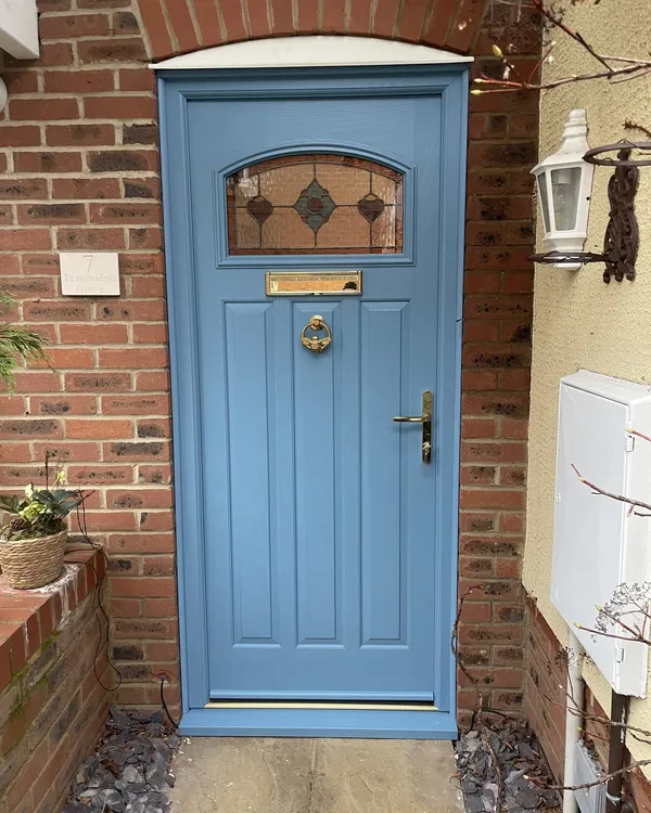 Mid blue cottage-style composite door with a stained glass top panel and brass knocker, handle and letterplate, under a brick arch.