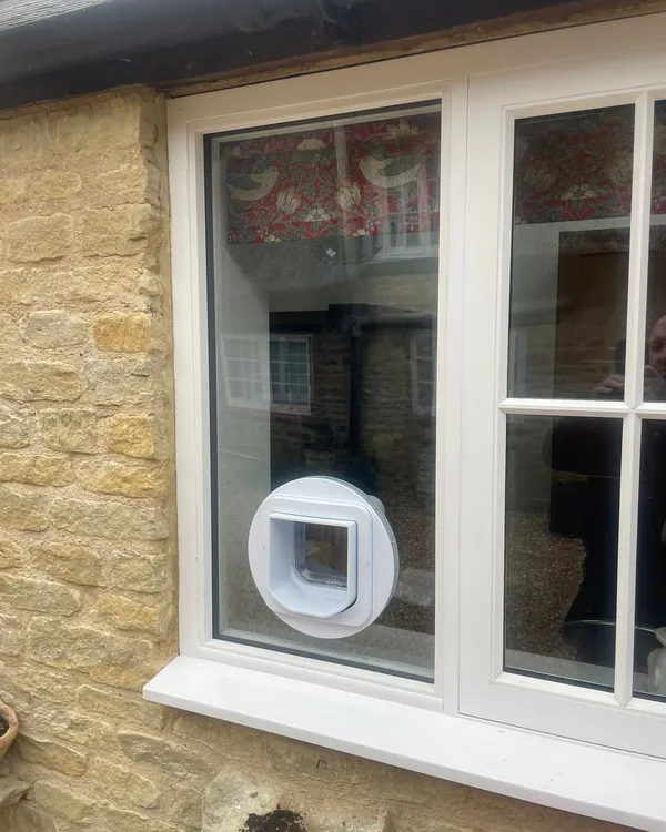White microchip cat flap fitted into the lower glass pane of a white window in a honey-coloured stone cottage.