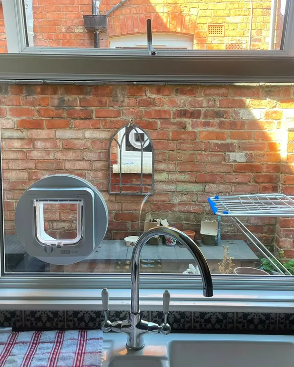 White SureFlap microchip cat flap set into the glass of a kitchen window above a Belfast sink, with a circular mounting adaptor.