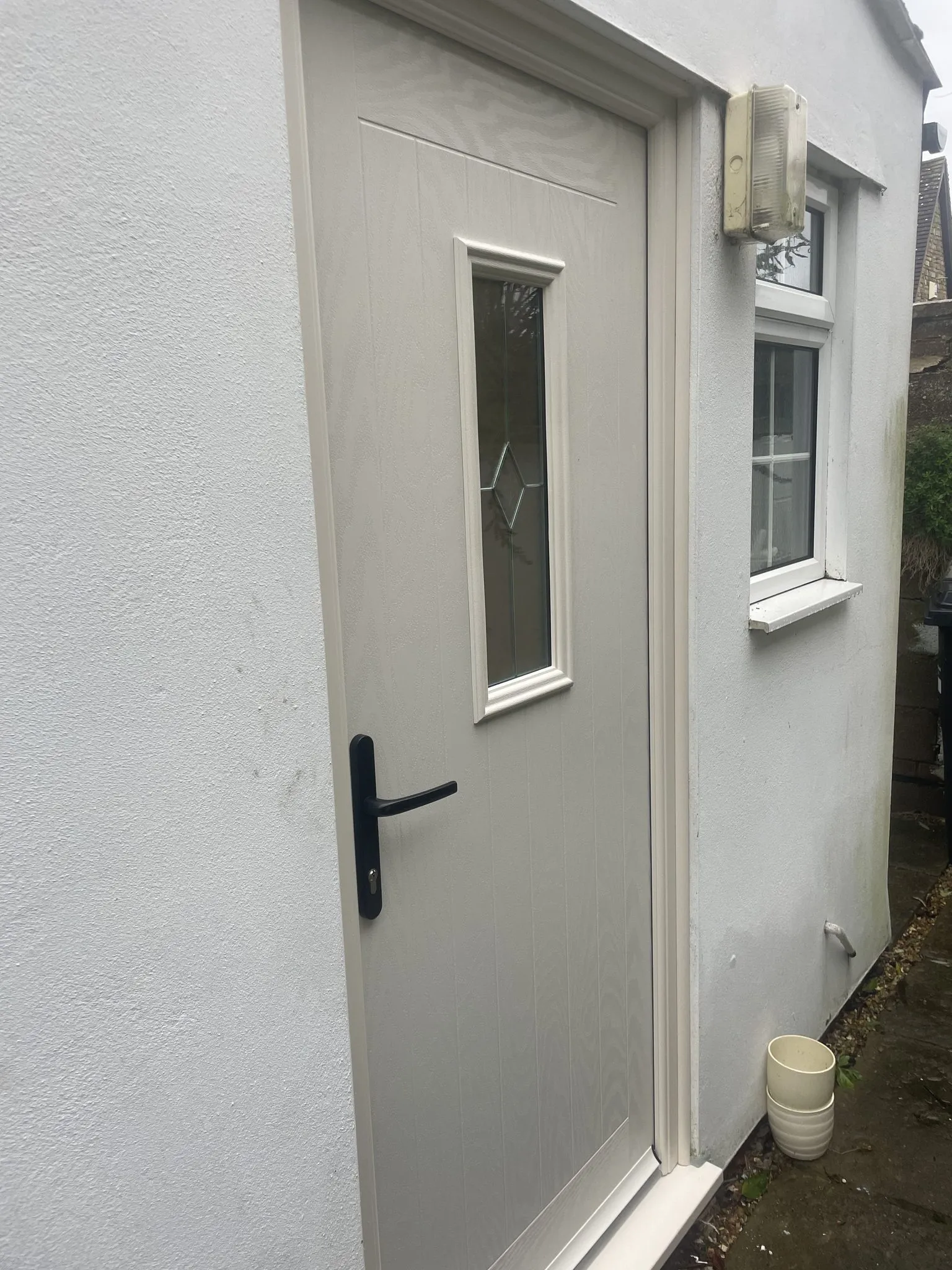 Cream composite door with a slim decorative glass panel and black lever handle, fitted in a white rendered wall.