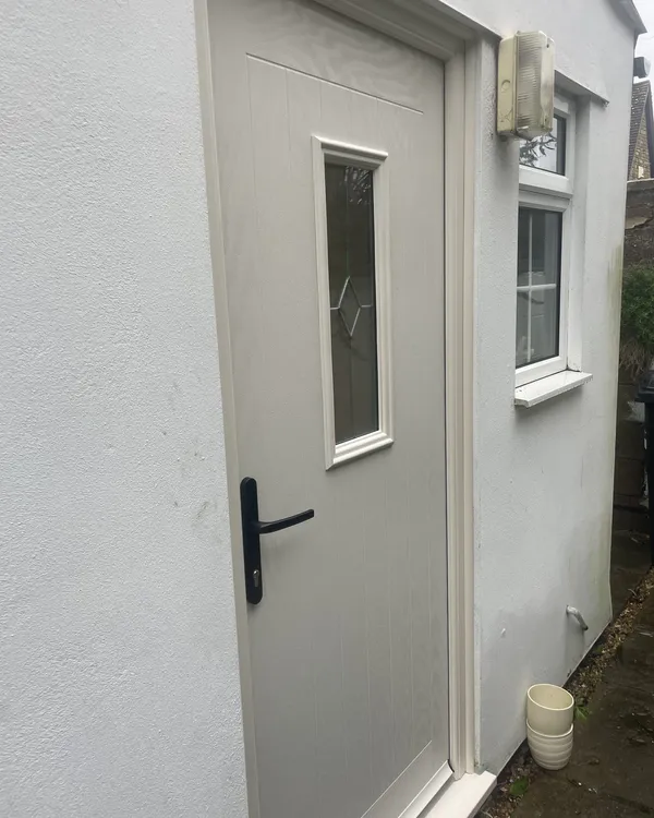 Cream composite door with a slim decorative glass panel and black lever handle, fitted in a white rendered wall.