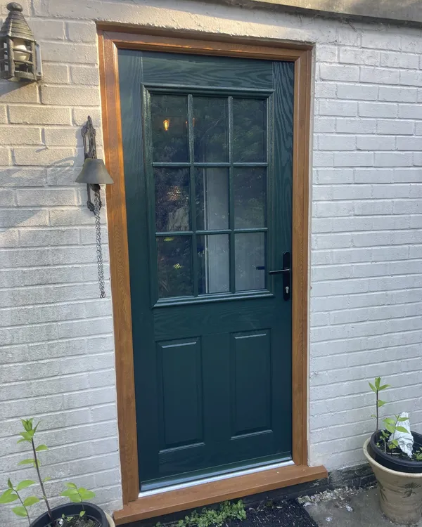 Dark green composite door with a Georgian-barred glazed top half, fitted in an oak-effect frame in a painted brick wall.