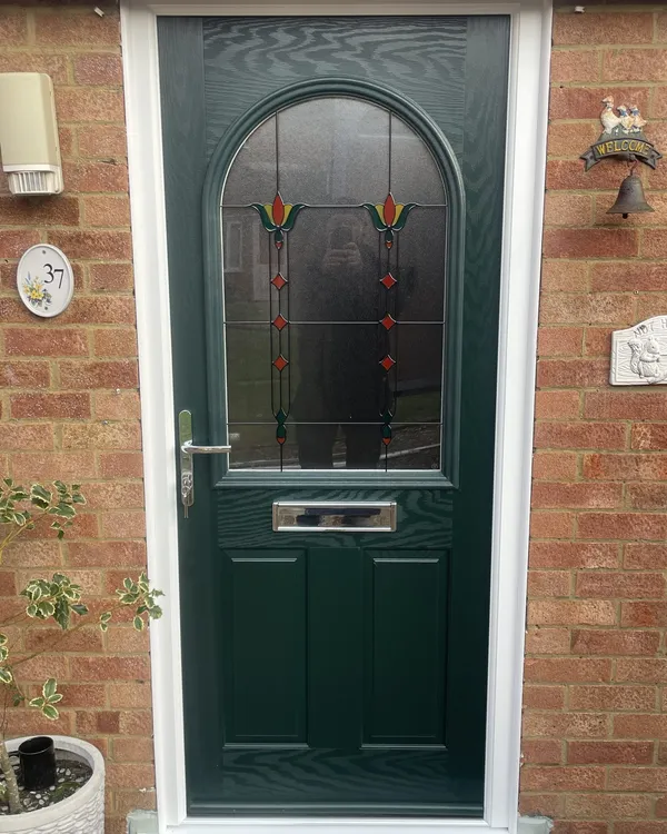 Dark green composite front door with a large arched glass panel featuring stained glass tulips, in a white frame on a brick house.