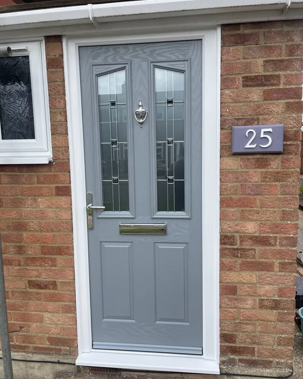 Mid grey composite front door with two decorative glazed panels and a chrome knocker, in a white frame on a red brick house.