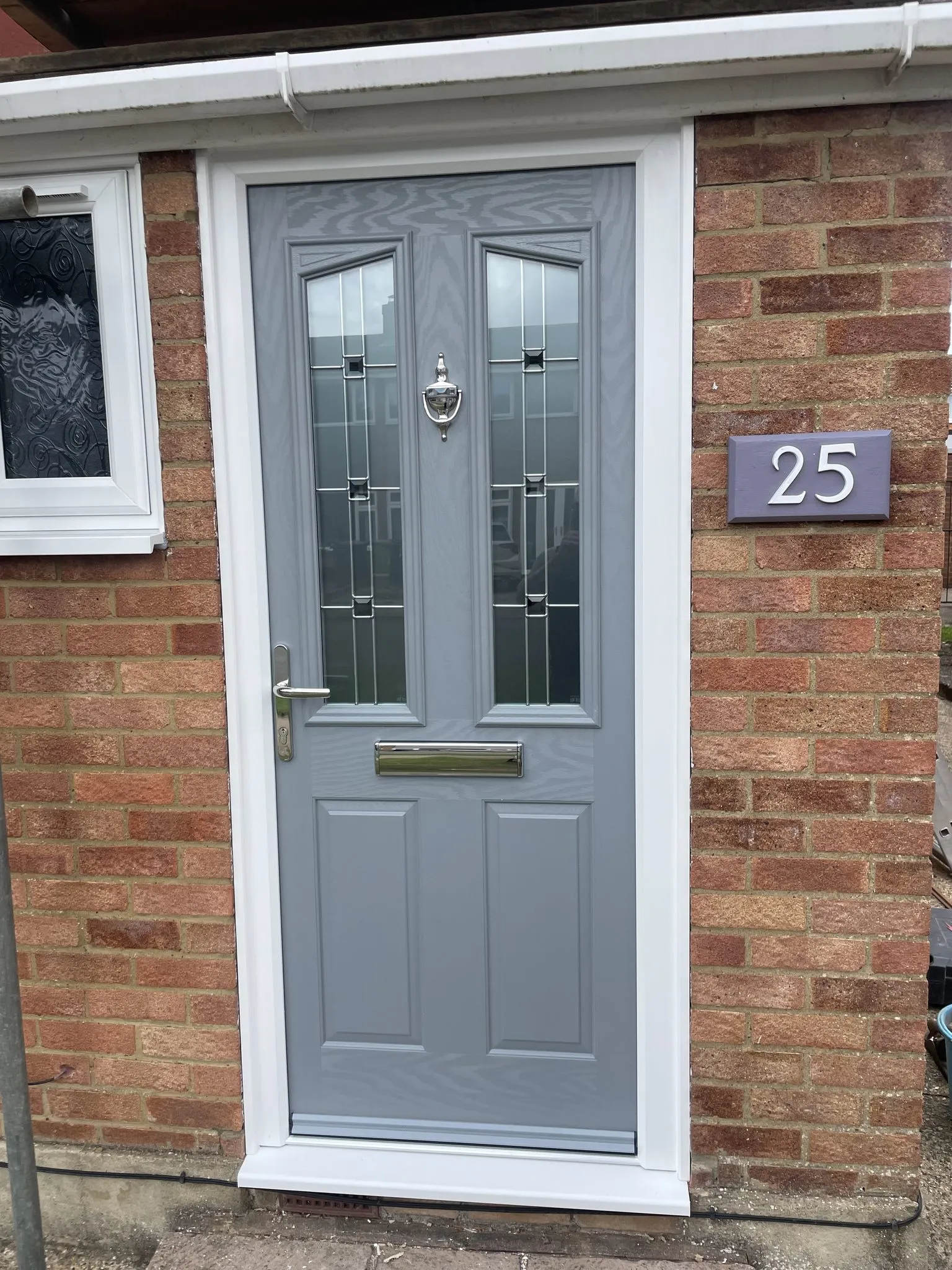 Mid grey composite front door with two decorative glazed panels and a chrome knocker, in a white frame on a red brick house.