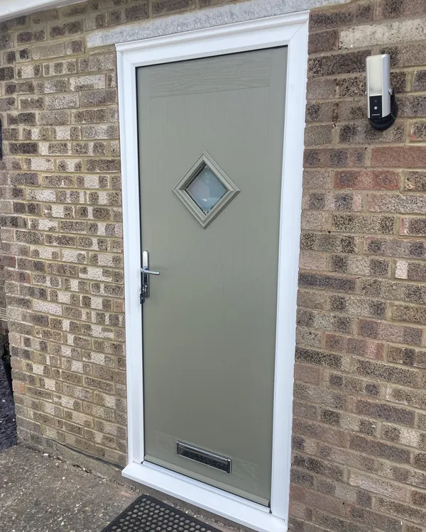 Grey-green composite door with a diamond-shaped glass panel and chrome furniture, in a white frame on a brick house.