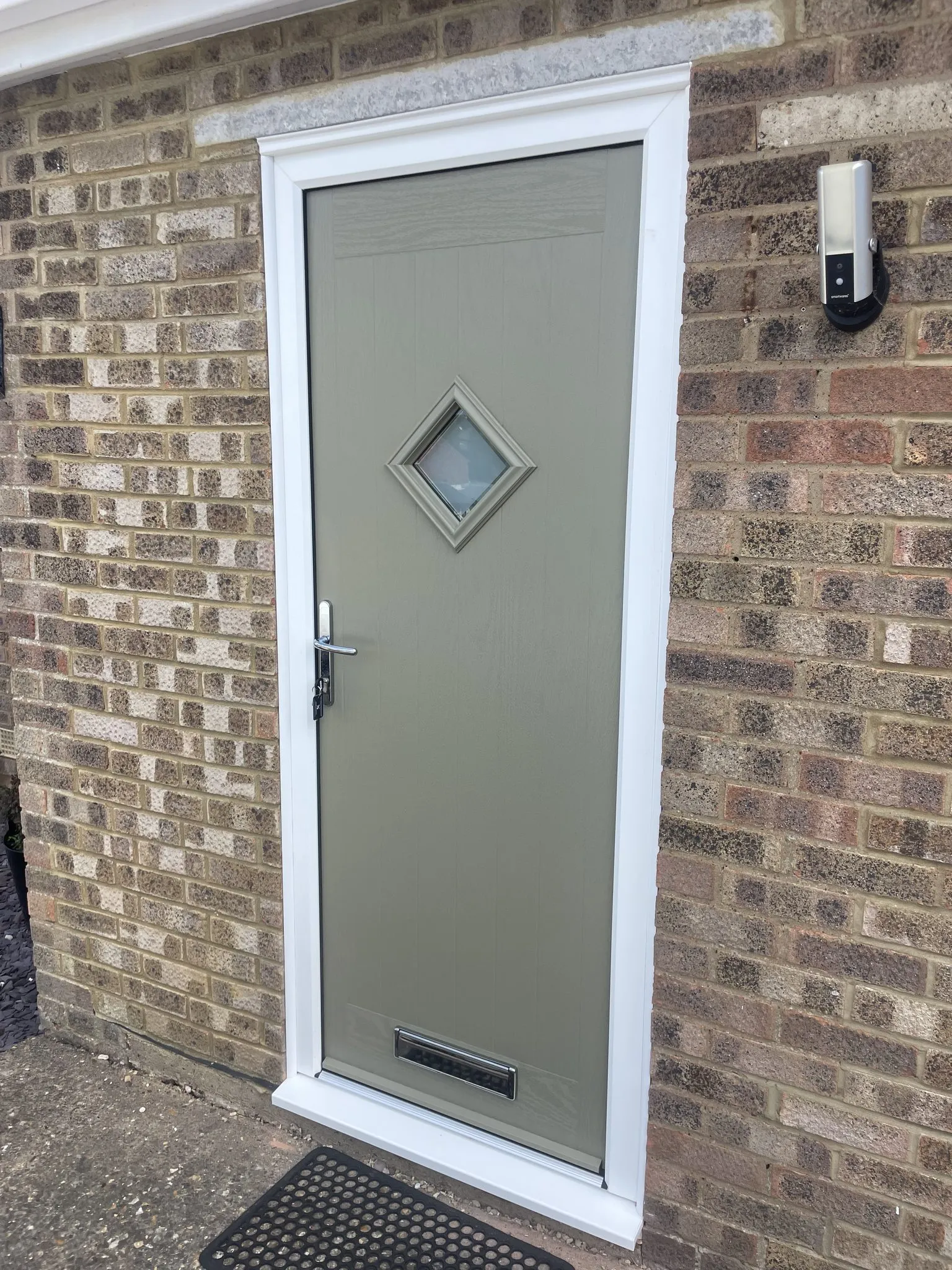 Grey-green composite door with a diamond-shaped glass panel and chrome furniture, in a white frame on a brick house.