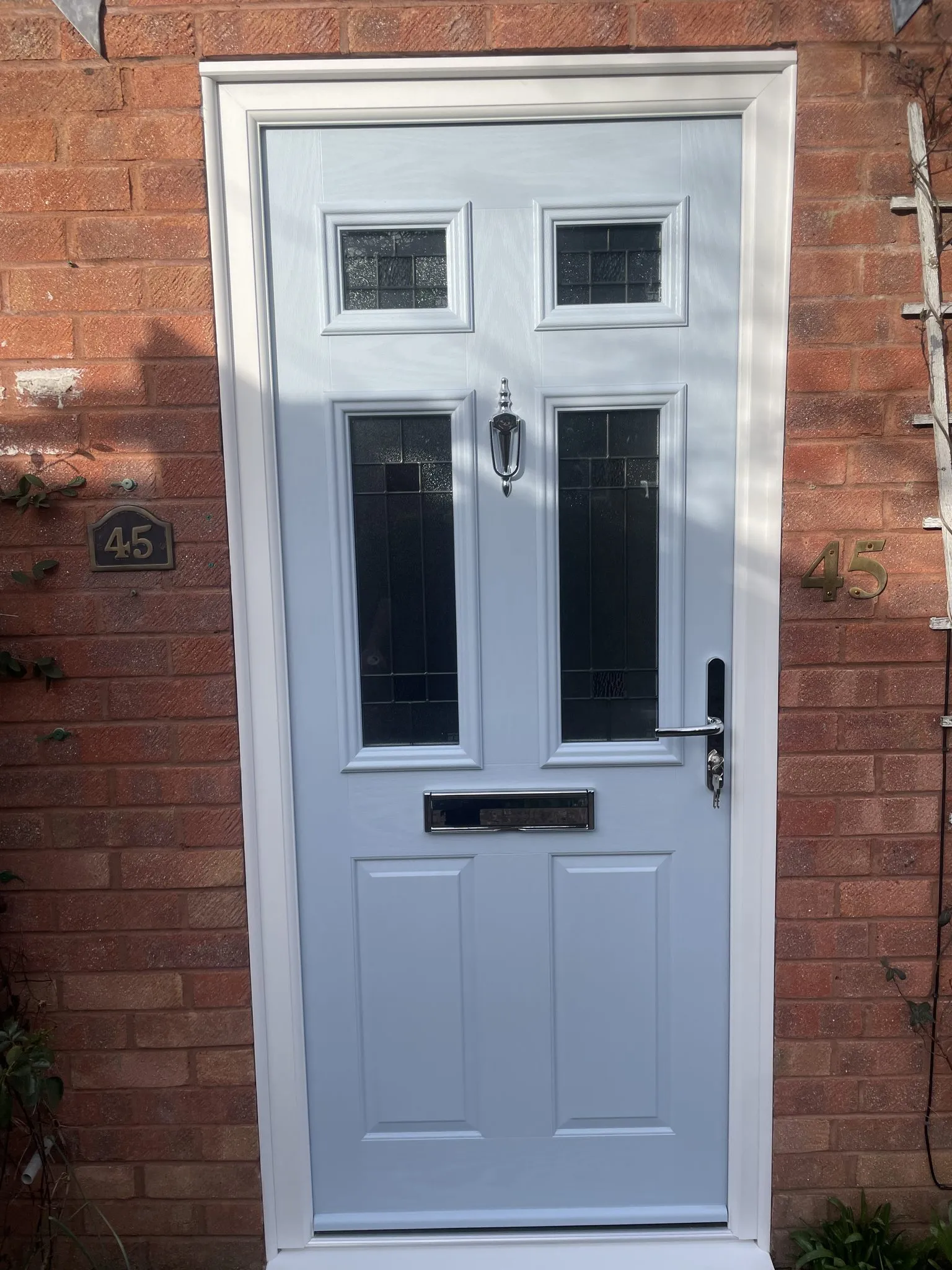 Light grey composite front door with four leaded glass panels and a chrome knocker, in a white frame on a red brick house.