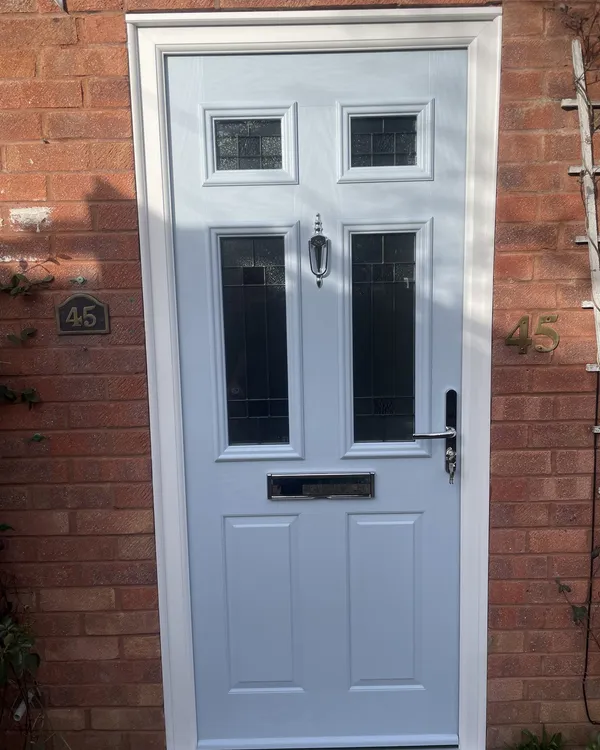 Light grey composite front door with four leaded glass panels and a chrome knocker, in a white frame on a red brick house.
