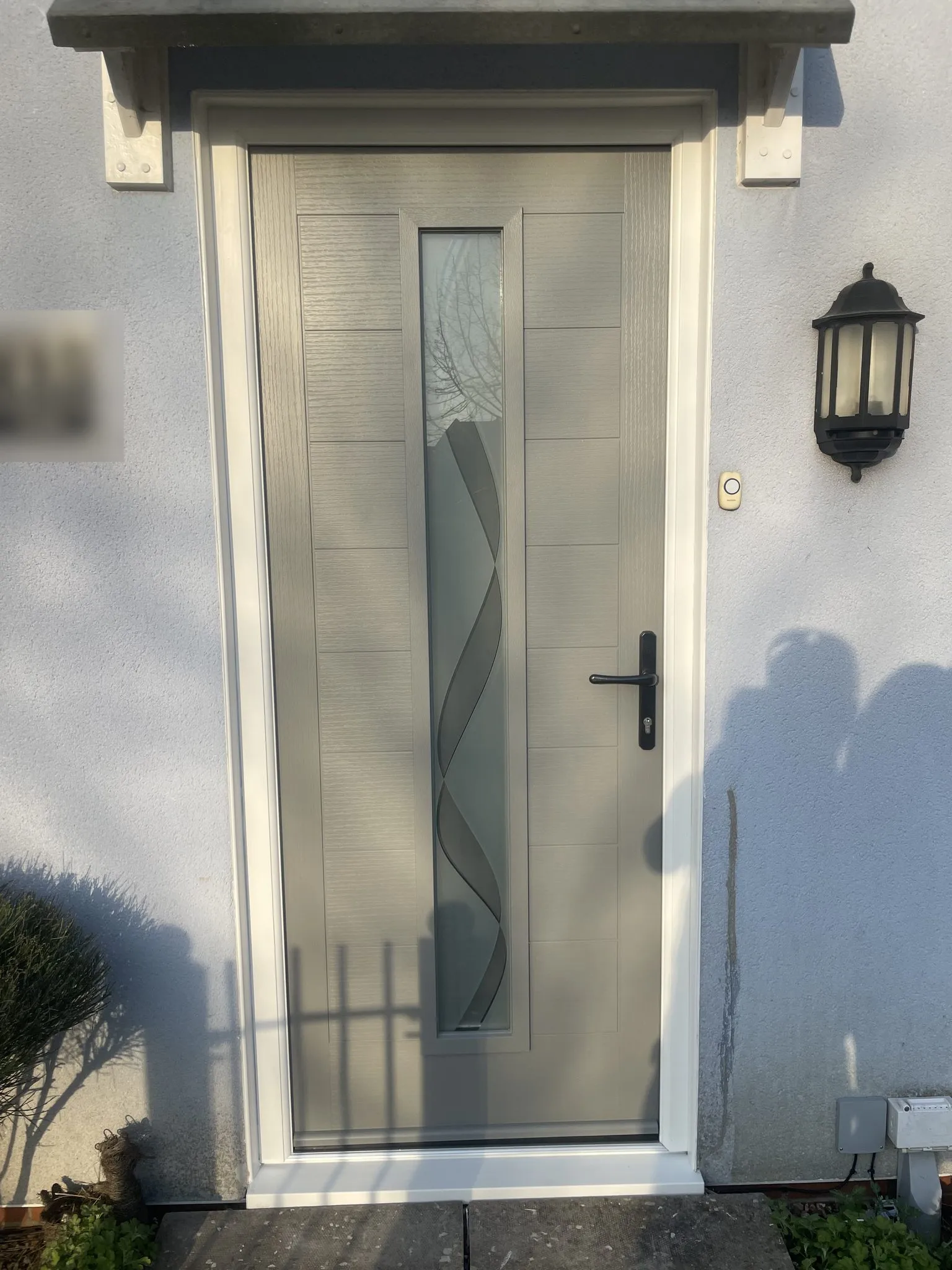 Modern light grey composite door with grooved panels, a tall decorative glass strip and black lever handle, under a canopy on a rendered house.