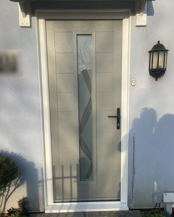 Modern light grey composite door with grooved panels, a tall decorative glass strip and black lever handle, under a canopy on a rendered house.