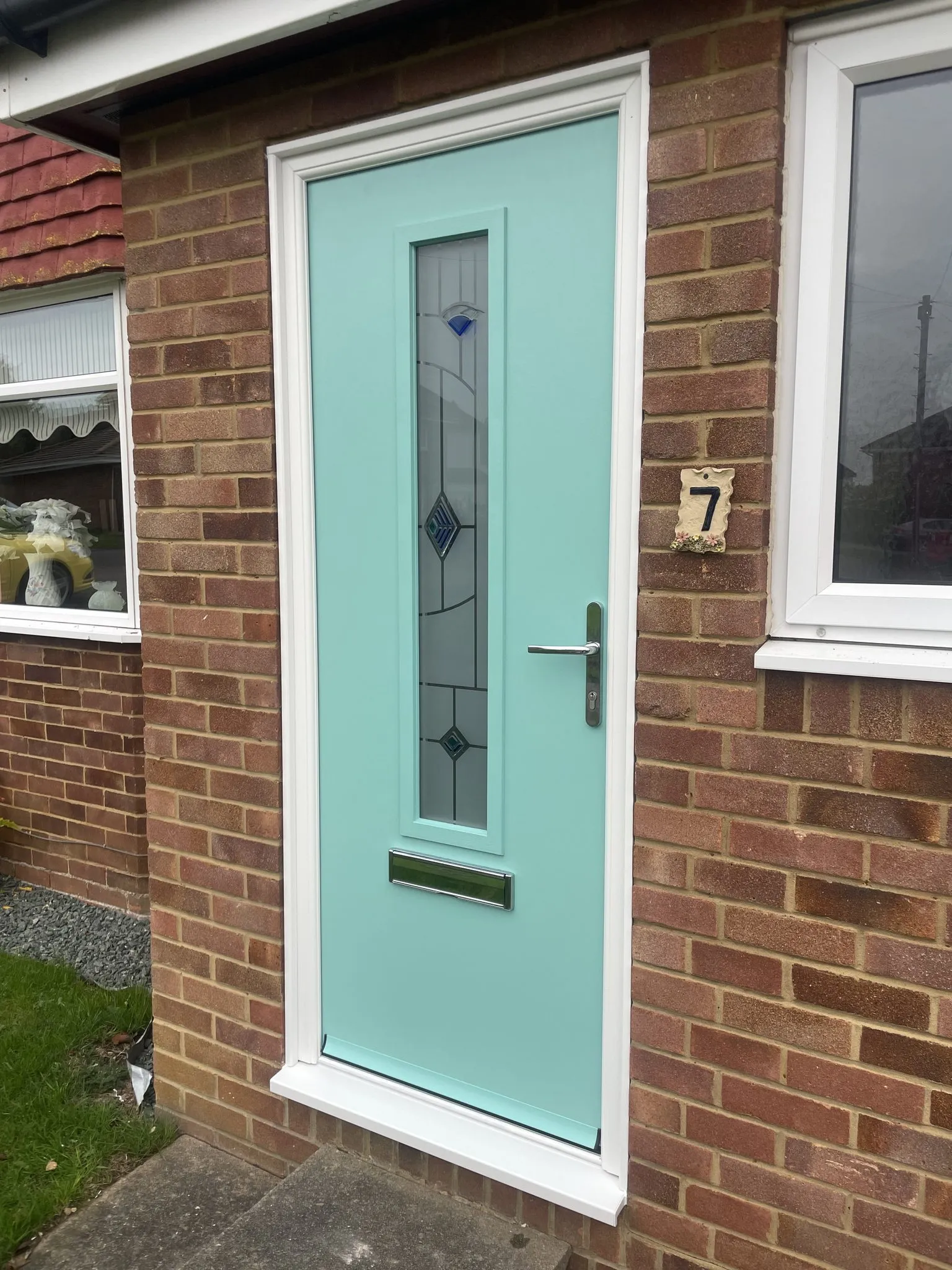 Mint green composite front door with a single tall decorative glass panel and chrome handle, in a white frame on a brick house.
