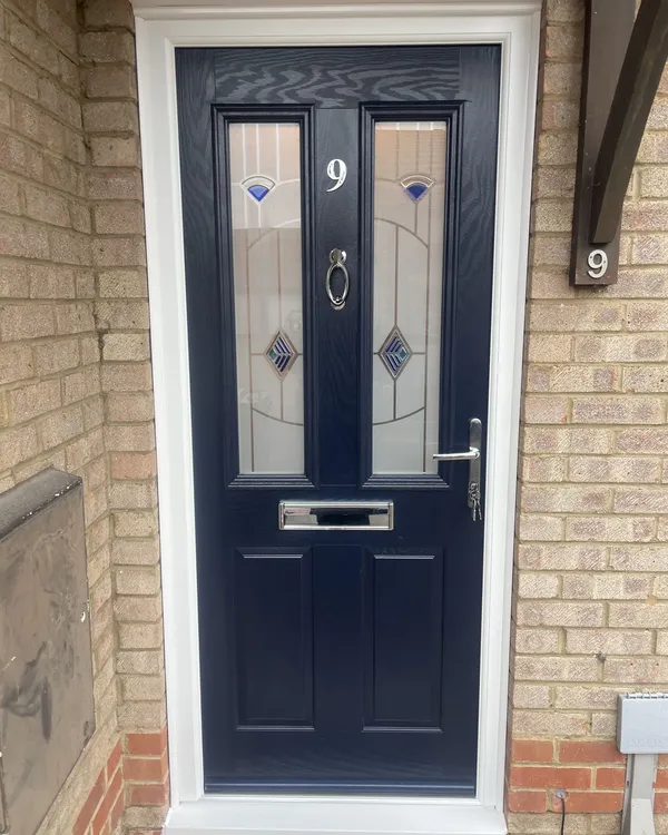 Navy blue composite front door with decorative glass panels featuring blue jewels and diamond leading, in a white frame on a buff brick house.