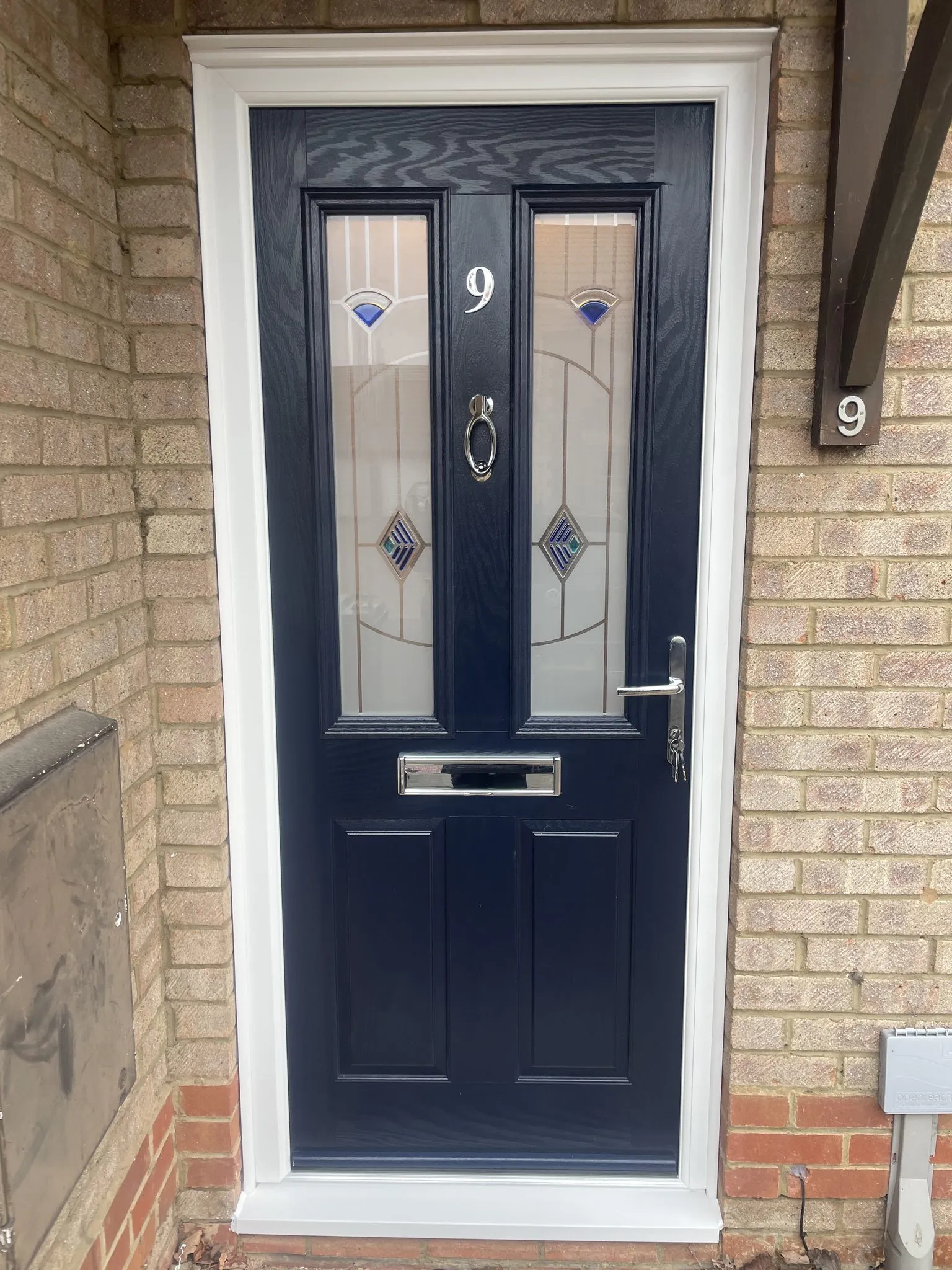 Navy blue composite front door with decorative glass panels featuring blue jewels and diamond leading, in a white frame on a buff brick house.
