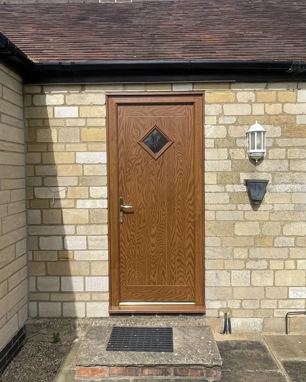 Golden oak woodgrain composite door with a diamond-shaped glass panel and brass handle, fitted in a stone wall.