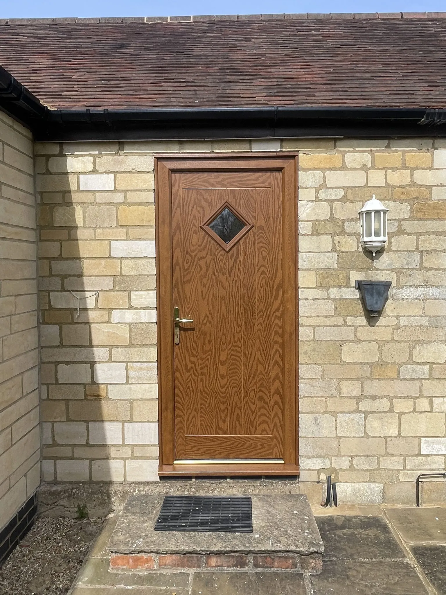Golden oak woodgrain composite door with a diamond-shaped glass panel and brass handle, fitted in a stone wall.