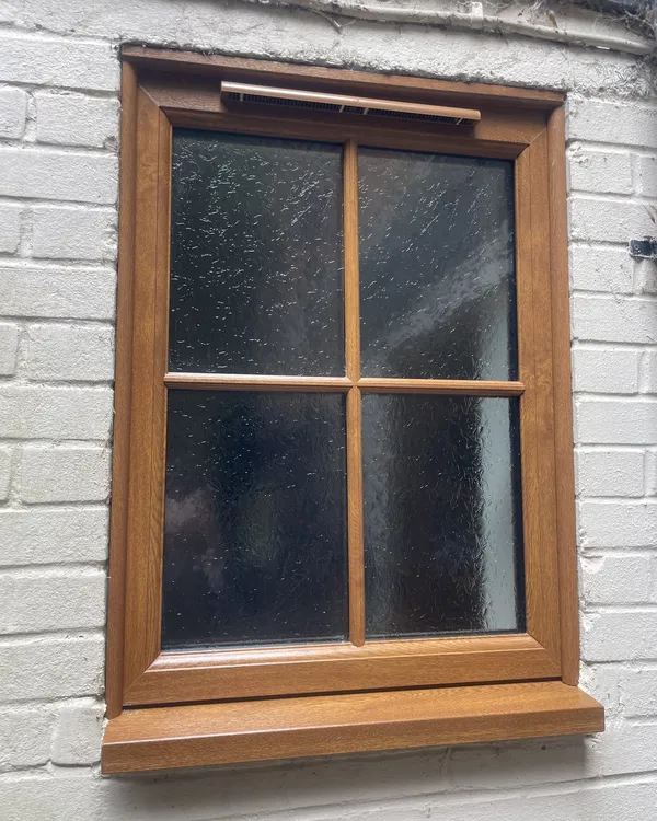 Golden oak woodgrain uPVC window with Georgian bars and obscure glass, fitted in a painted brick wall.