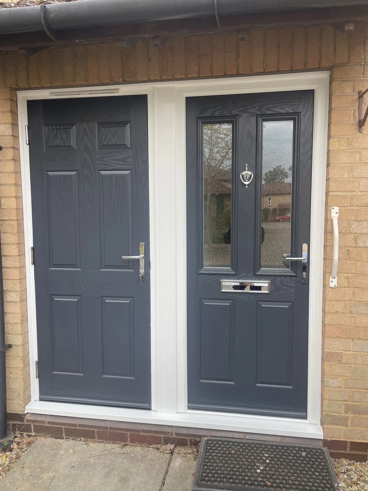 Two anthracite grey composite doors side by side in a shared white frame, one with glazed panels and one solid panelled.
