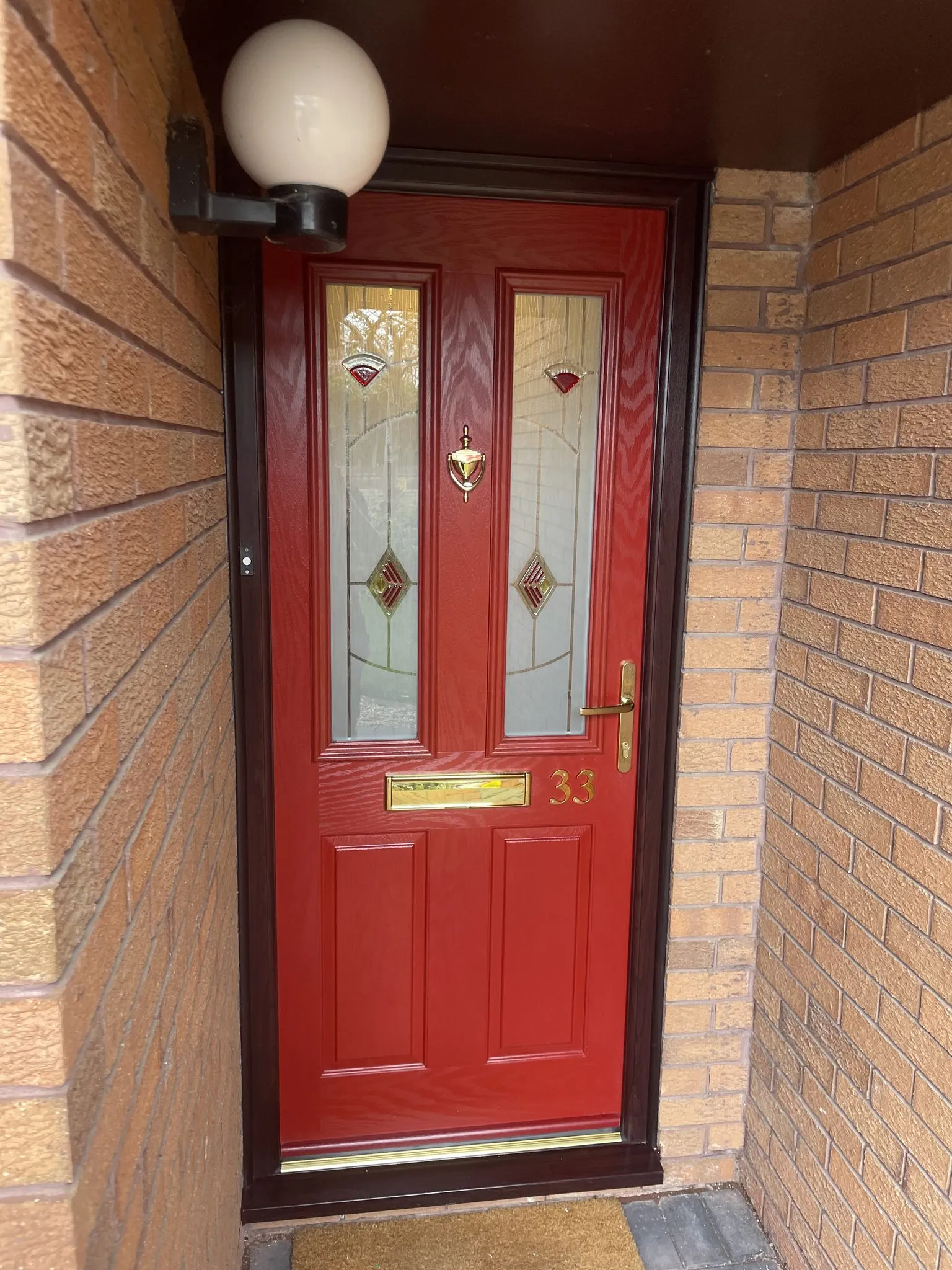 Red composite front door with decorative glass panels and a brass knocker, handle and letterplate, in a rosewood-effect frame inside a brick porch.