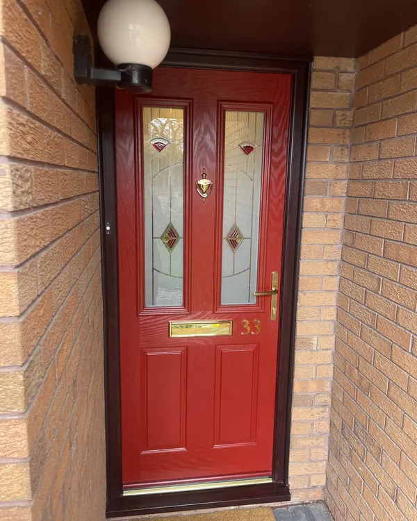 Red composite front door with decorative glass panels and a brass knocker, handle and letterplate, in a rosewood-effect frame inside a brick porch.