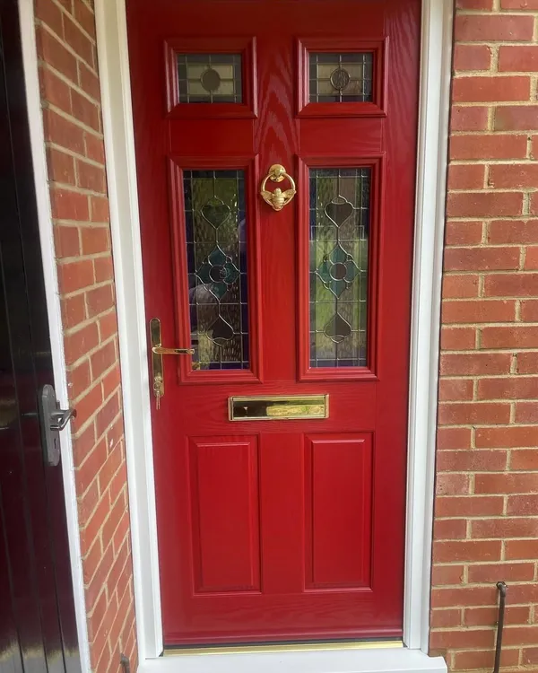 Red composite front door with stained glass panels, a brass knocker, handle and letterplate, in a white frame.