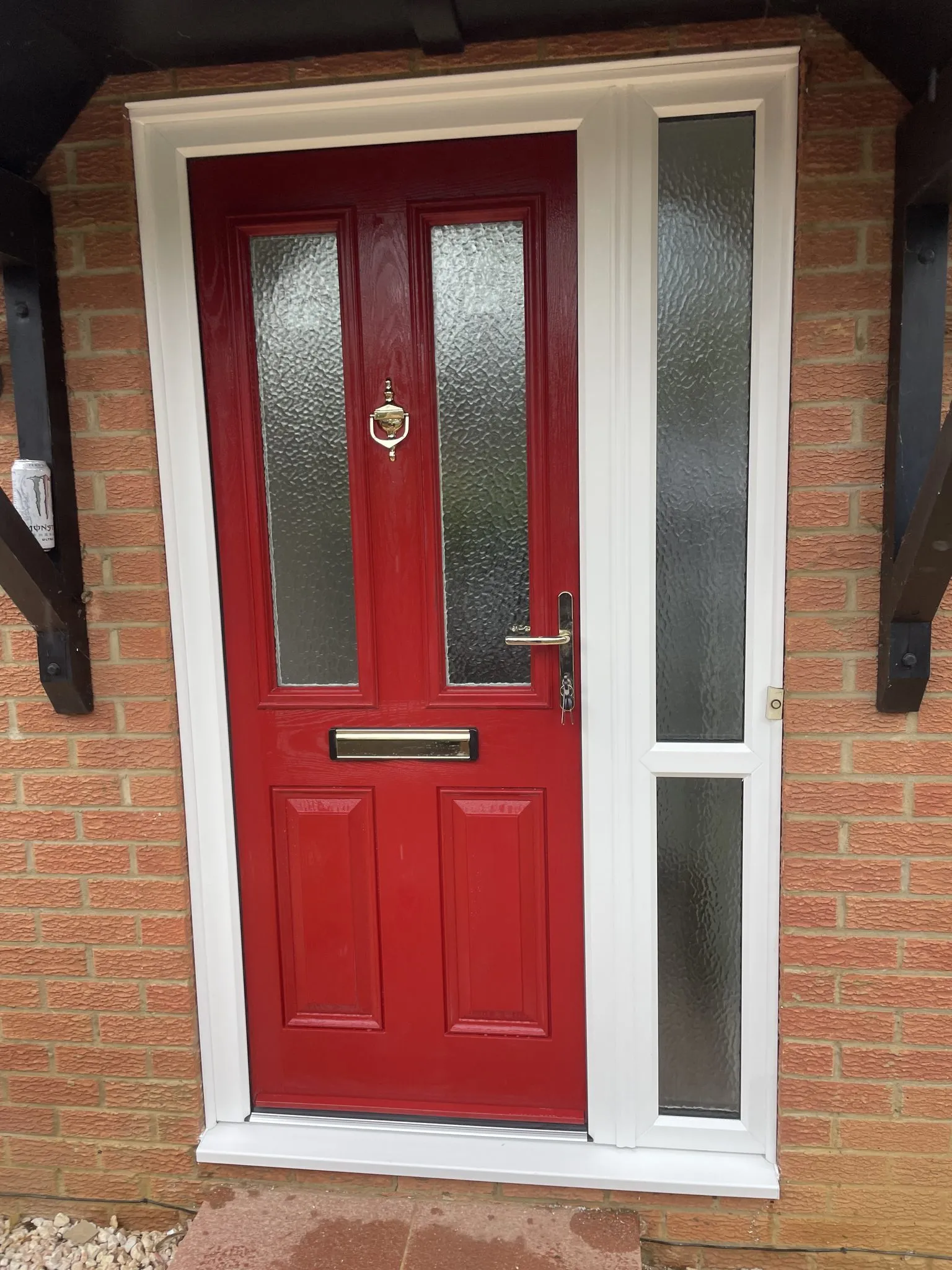 Red composite front door with obscure glass panels and chrome furniture, in a white frame with a glazed side panel beneath a timber canopy.