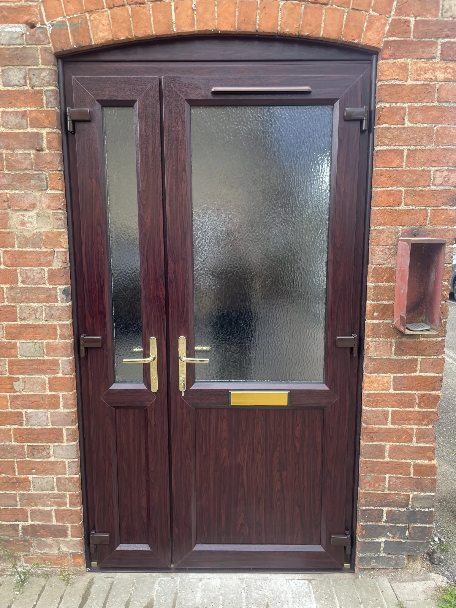 Rosewood-effect uPVC double doors with obscure glass and brass handles, fitted under a curved brick arch.